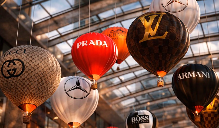 Кинетическая инсталляция «Shopping Vortex» — подвесная композиция из брендированных воздушных шаров Prada, Louis Vuitton, Gucci, Armani, Boss и Mercedes под стеклянным куполом, с тёплой подсветкой, создающей эффект роскошного вихря покупок.