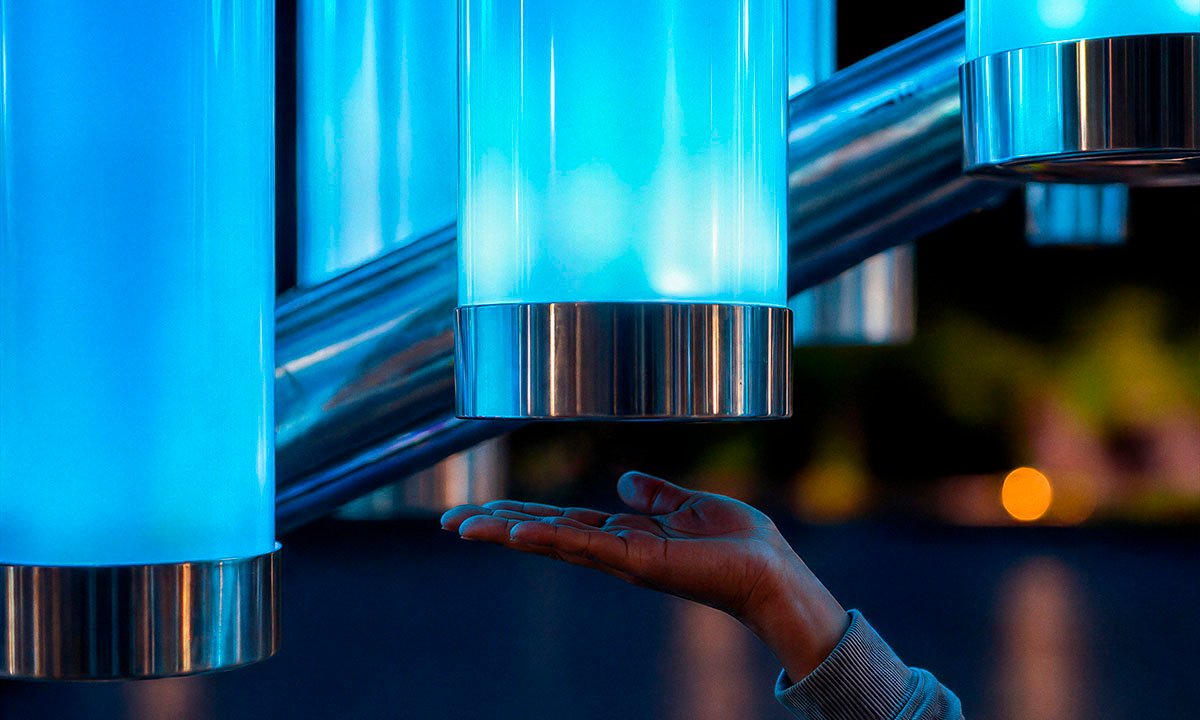 Close-up of the interactive installation “Magic Streams” — an illuminated cylinder made of matte polycarbonate with metal elements, responding to the approach of a hand; the soft blue glow intensifies upon interaction, creating a responsive light effect.