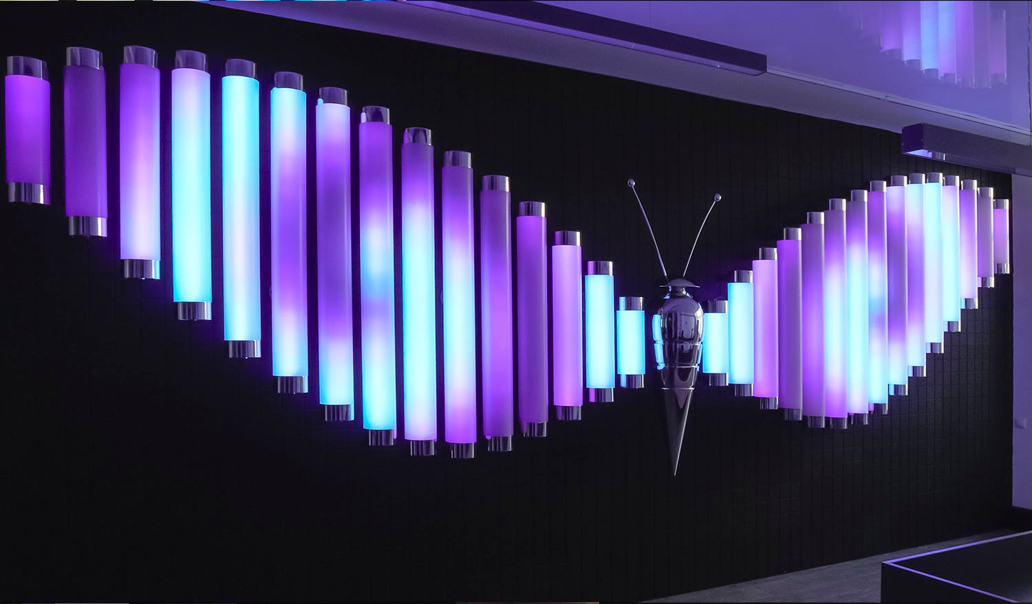Interactive Butterfly light installation with wings made of glowing violet and cyan tubes and a metallic butterfly sculpture at the center.