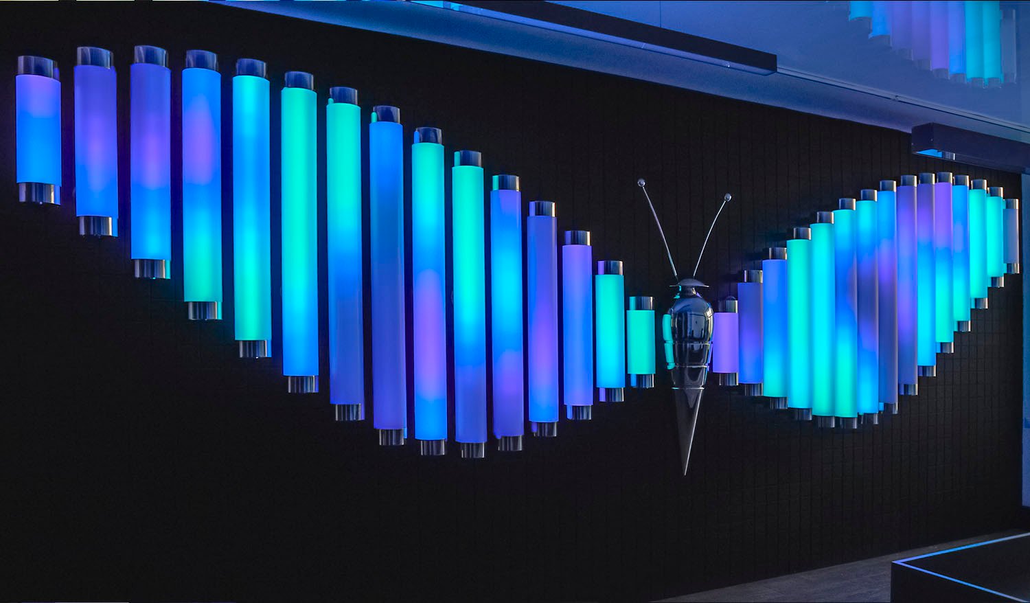 Interactive Butterfly light installation with wings made of glowing blue, turquoise, and violet tubes and a metallic butterfly sculpture at the center.